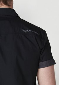 Camisa de manga corta negra hecha de una tela ligera con un puño enrollado gris, con "KOROSHI BRAND" y coordenadas geográficas impresas en la parte posterior.