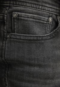 Zwarte spijkerbroek van denim toont een close-up van een voorzak met zilveren details. De stof vertoont een lichte textuur en nette stiknaden.