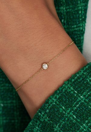 Goldene Kettenarmband mit einem zarten Design, das einen einzelnen runden klaren Stein in der Mitte hat. Am gebräunten Handgelenk getragen.