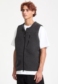 Grå fleecevest med lynlåslommer, høj krave og en struktureret overflade. Bæres over en hvid t-shirt med en afslappet pasform.