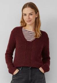 Frau mit hellbraunem Haar trägt einen weinroten Strickpullover über einem weiß und weinrot gestreiften Oberteil, die Hände in den Taschen von schwarzen Jeans, vor einem schlichten Hintergrund.