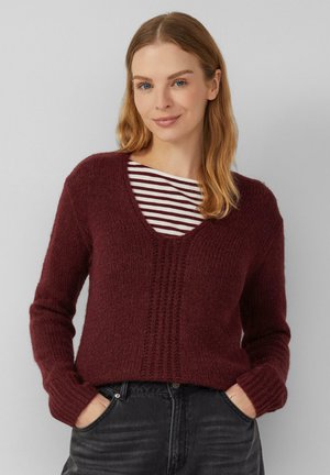 Femme aux cheveux châtain clair portant un pull en tricot bordeaux sur un haut rayé blanc et bordeaux, les mains dans les poches d'un jean noir, sur fond uni.