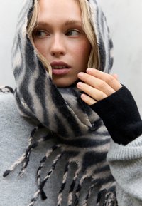 Écharpe en laine rayée noire et beige avec des franges, enroulée autour du visage, associée à un pull gris clair et des mitaines noires.