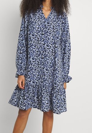 Kvinde iført en løs, langærmet, knælængde kjole med flæsekant og et blåt blomsterprint på en mørk baggrund.