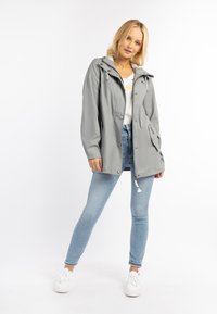 Giacca impermeabile grigia chiara con cappuccio, bottoni a scatto e tasche laterali. Abbinata a jeans aderenti blu chiaro e scarpe da ginnastica bianche.