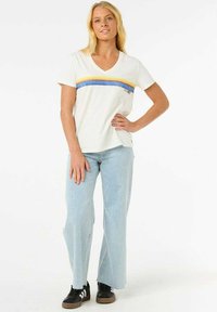 Weißes Baumwoll-T-Shirt mit V-Ausschnitt, mit horizontalen Streifen in Gelb, Orange und Blau. Getragen mit hellblauen Schlagjeans und schwarzen Sneakers.