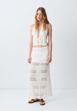Jeune femme aux longs cheveux portant un crop top blanc sans manches à col halter et une longue jupe blanche en dentelle associée à des sandales marron.