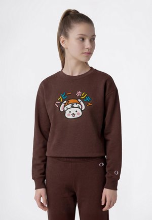 Ung kvinde med hestehale iført en brun sweatshirt og bukser, sweatshirtens motiv er en tegneseriehund med farverig japansk tekst ovenover.