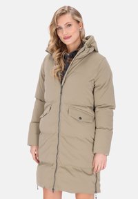 Vrouw in een beige gewatteerde lange jas met een capuchon, voorrits en klepzakken over een geruit shirt, lachend met golvend blond haar.