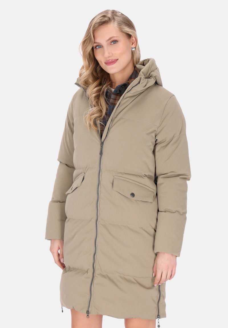 Vrouw in een beige gewatteerde lange jas met een capuchon, voorrits en klepzakken over een geruit shirt, lachend met golvend blond haar.