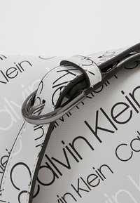 Vit läderväska med ett upprepande svart "Calvin Klein" logomönster, med en elegant design, justerbar rem och metalliska detaljer.