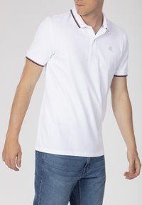 Polo shirt blanc en coton, doté d'un col côtelé avec des rayures rouges et bleues et d'un logo brodé sur la poitrine. Manches courtes.
