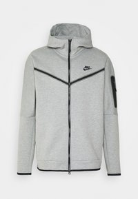 Felpa grigia con zip frontale, dettagli neri, logo Nike sul petto e tasca con zip sul braccio sinistro. Tessuto morbido e texturizzato.