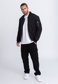 Giacca bomber nera con zip frontale, tasche laterali e fodera rossa, indossata sopra una t-shirt bianca e pantaloni cargo neri, completata da sneakers bianche.