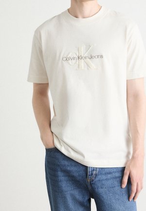 Camiseta de algodón color beige claro con un logo texturizado de "Calvin Klein Jeans" en colores contrastantes. Mangas cortas, cuello redondo, corte relajado.