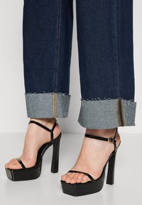 Jeans de pierna ancha de color azul oscuro con puños deshilachados y detalles de denim en contraste, combinados con sandalias de tacón alto de charol negro con tiras finas.