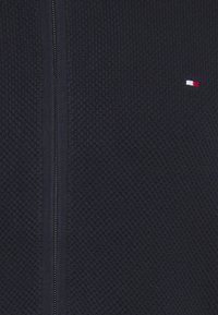 Chaqueta de punto con cremallera en color azul marino, con un patrón texturizado, que presenta una cremallera plateada y un pequeño logo en rojo, blanco y azul en la parte superior izquierda.