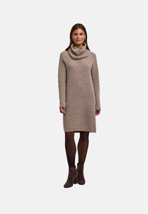 Brede gebreide trui jurk in beige met een col nek, lange mouwen en een relaxte pasvorm, gecombineerd met donkere panty’s en bruine enkellaarsjes.