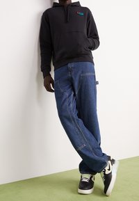 Svart hoodie med framficka och liten logga, kombinerad med blåa baggy jeans med sidostickning och två bakfickor. Svarta sneakers med vita och gröna detaljer.