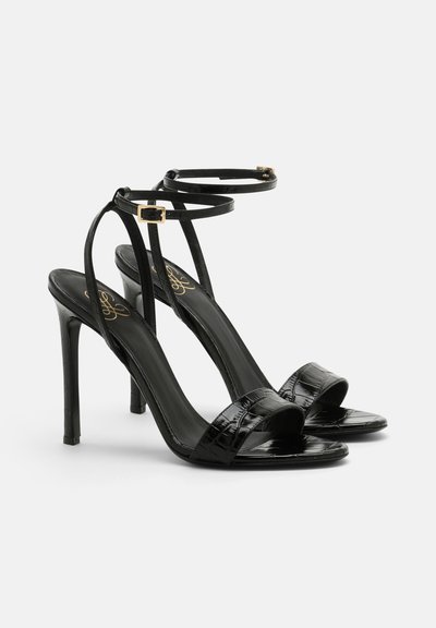 Sandalias de tacón alto negras con textura de cocodrilo brillante, que cuentan con finas correas en el tobillo y una sola banda frontal ancha. Detalles en dorado.