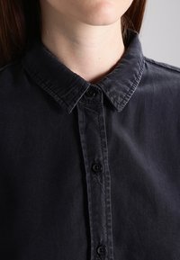 Camicia nera con colletto classico, realizzata in un tessuto morbido. Presenta una leggera texture e sei bottoni sulla parte anteriore.