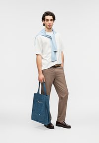 Τσάντα tote από τζιν με μπροστινή τσέπη με φερμουάρ, μπλε χρώμα, ρυθμιζόμενους ιμάντες και υφή επιφάνειας. Το μοντέλο φοράει ανοιχτόχρωμο πουκάμισο και καφέ παντελόνι.