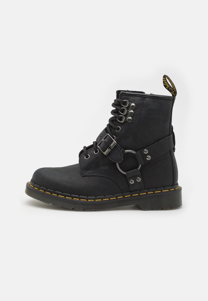 Botte de combat en cuir noir avec devant lacé, sangle à boucle métallique, détail d’anneau rond argenté et semelle cousue jaune.