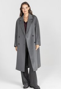 Szary podwójnie zapinany płaszcz z dużymi guzikami, szerokimi klapami i luźnym krojem, wykonany z miękkiego materiału, połączony z ciemnymi spodniami o szerokich nogawkach.
