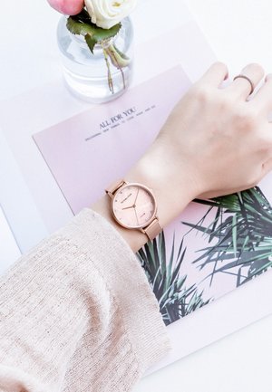 Poignet avec montre en or rose et bague en argent reposant sur des magazines aux couvertures florales et feuilles de palmier, à côté d'un petit vase contenant des fleurs roses et blanches.