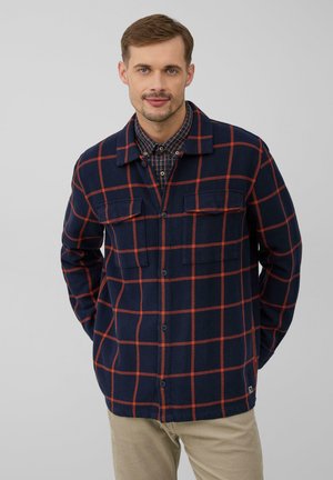 Man in een marineblauw en rood geruit overhemd met knopen over een overhemd, poseert met handen achter de rug tegen een grijze achtergrond.