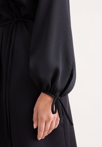 Schwarzes, langärmeliges Kleid mit gerafften Bündchen und Schleifenakzenten. Weicher, glatter Stoff; besitzt einen subtilen Glanz für mehr Struktur.