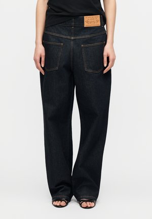 Personne portant un jean denim bleu foncé ample avec de grandes poches arrière et une pièce en cuir sur la ceinture, assorti à des sandales noires et un haut noir.