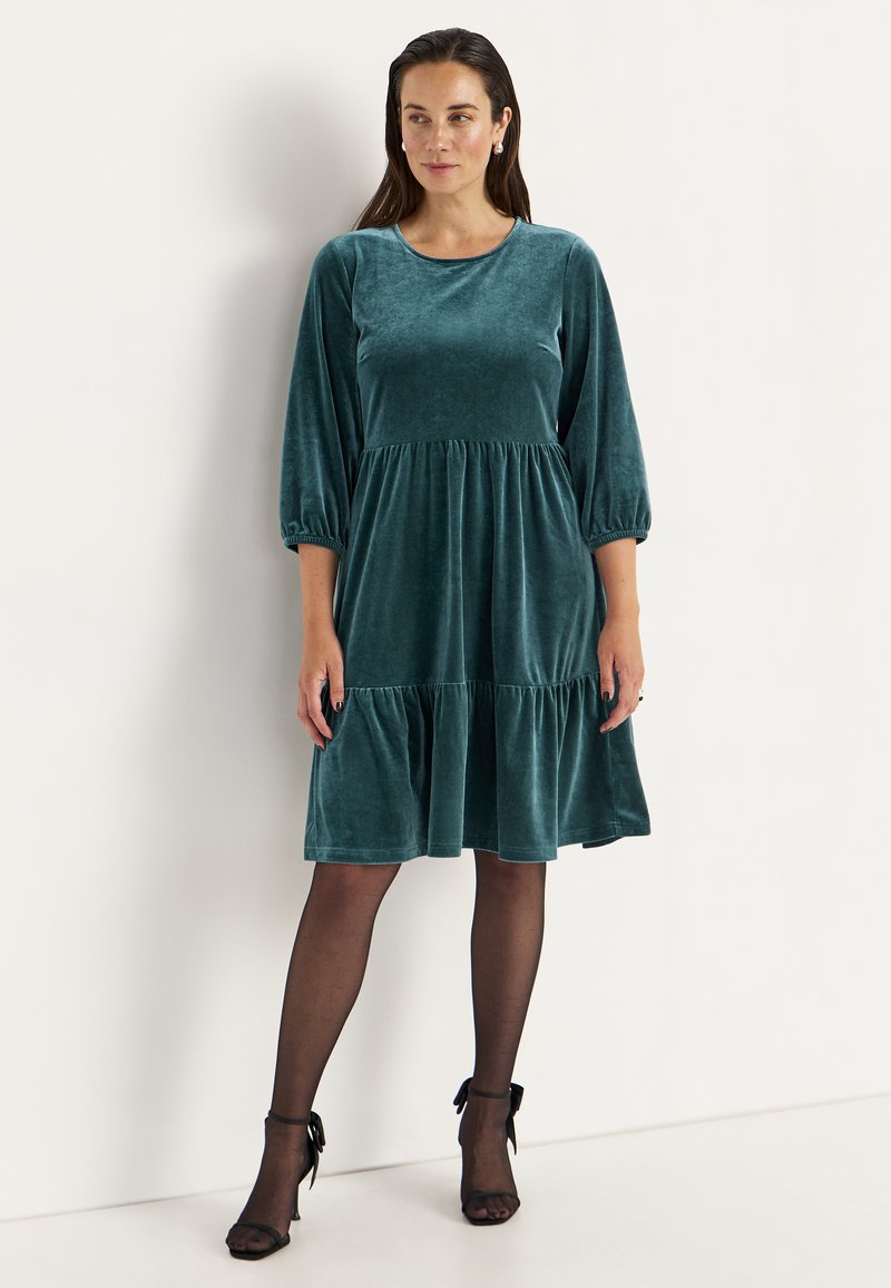 Teal fluwelen jurk met een ronde halslijn, pofmouwen en een gelaagde rok. Gemêleerd stof met een zachte glans en een gerimpelde taille-detail.