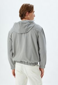 Chaqueta con capucha de color gris claro, con cintura y puños elásticos, presenta un diseño sencillo y una textura suave, llevada sobre pantalones blancos.