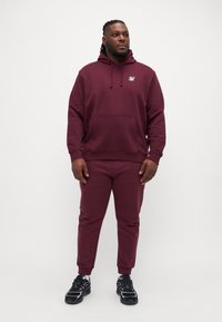 Bordeaux hettegenser med framlomme og snor, kombinert med matchende joggebukser. Antrekket har en liten logo på brystet og joggesko.
