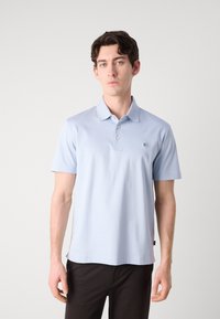 Polo de cor azul-claro, feito de tecido macio, com um colarinho clássico, três botões e um pequeno logotipo no peito. Mangas curtas.