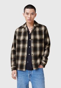 Ternet flannel skjorte i brun, creme og sort. Har en spids krave og to brystlommer. Bæres over en mørk skjorte med knapper.