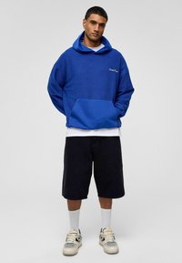Blauwe fleece hoodie met een buidelzak, witte geborduurde logo, gecombineerd met zwarte shorts en witte sokken, gedragen met tan sneakers.