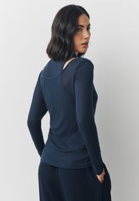 Maglia a maniche lunghe color blu navy con silhouette aderente, caratterizzata da ritagli trasparenti sulla schiena e tessuto morbido e strutturato. Abbinata a pantaloni scuri.