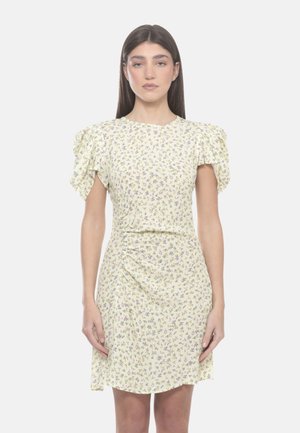 Donna che indossa un vestito floreale giallo chiaro con maniche corte a sbuffo e dettagli arricciati, in piedi davanti a uno sfondo bianco semplice.