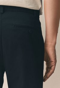 Pantalon noir à texture lisse, avec une poche arrière fermée par un bouton et un discret détail de double couture.