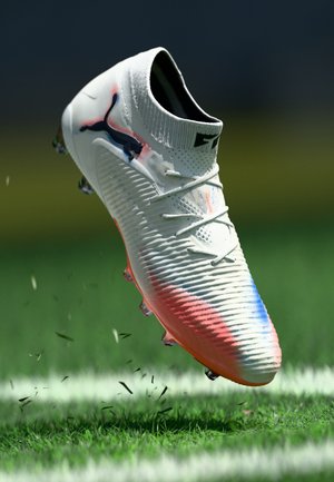 White soccer cleat with black Puma logo, red and blue accents, mid-air above grass field with blades scattering.