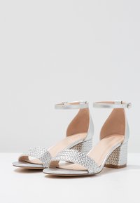 Silver open-toe sandals with a woven metallic strap and a textured block heel. Ankle strap with buckle adds support. Smooth inner lining.