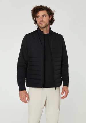 Gilet rembourré noir avec un design matelassé, col haut et poignets côtelés, porté sur un sweat-shirt noir, associé à un pantalon de couleur claire.