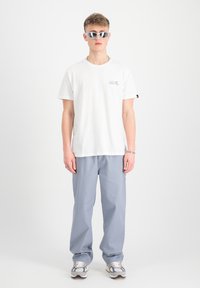 Camiseta de algodón blanca con el logo de "ALPHA INDUSTRIES" en azul y rojo, combinada con pantalones de algodón gris claro y zapatillas gris. Gafas de sol blancas.