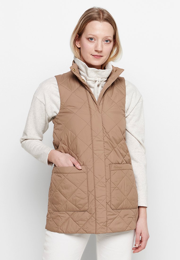 Woman wearing beige quilted sleeveless vest over light gray long-sleeve top with high collar, standing with one hand in pocket.