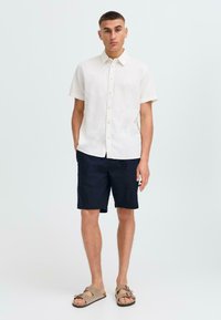 Camisa de lino de manga corta en color beige claro con cuello abotonado, combinada con pantalones cortos navy y sandalias marrones con dos correas ajustables.