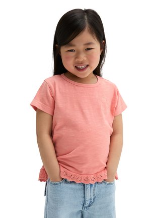 Lächelndes junges Mädchen mit langen dunklen Haaren, das ein rosa kurzärmeliges Shirt mit Spitzenbesatz und hellblaue Jeans trägt, Hände in den Taschen.