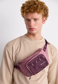 Nuori mies, jolla on kihara hiuksia, on pukeutunut beigeen huppariin ja vaaleanpunaiseen sekä viininpunaiseen olkalaukkuun, jossa on Columbian logo rinnalla, valkoista taustaa vasten.