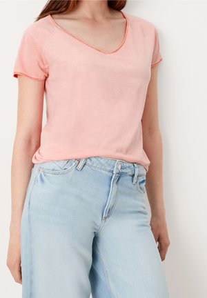 Kvinde iført en lyserød kortærmet V-hals skjorte, stoppet ned i lyseblå højtsiddende denimjeans, står op ad en hvid væg.